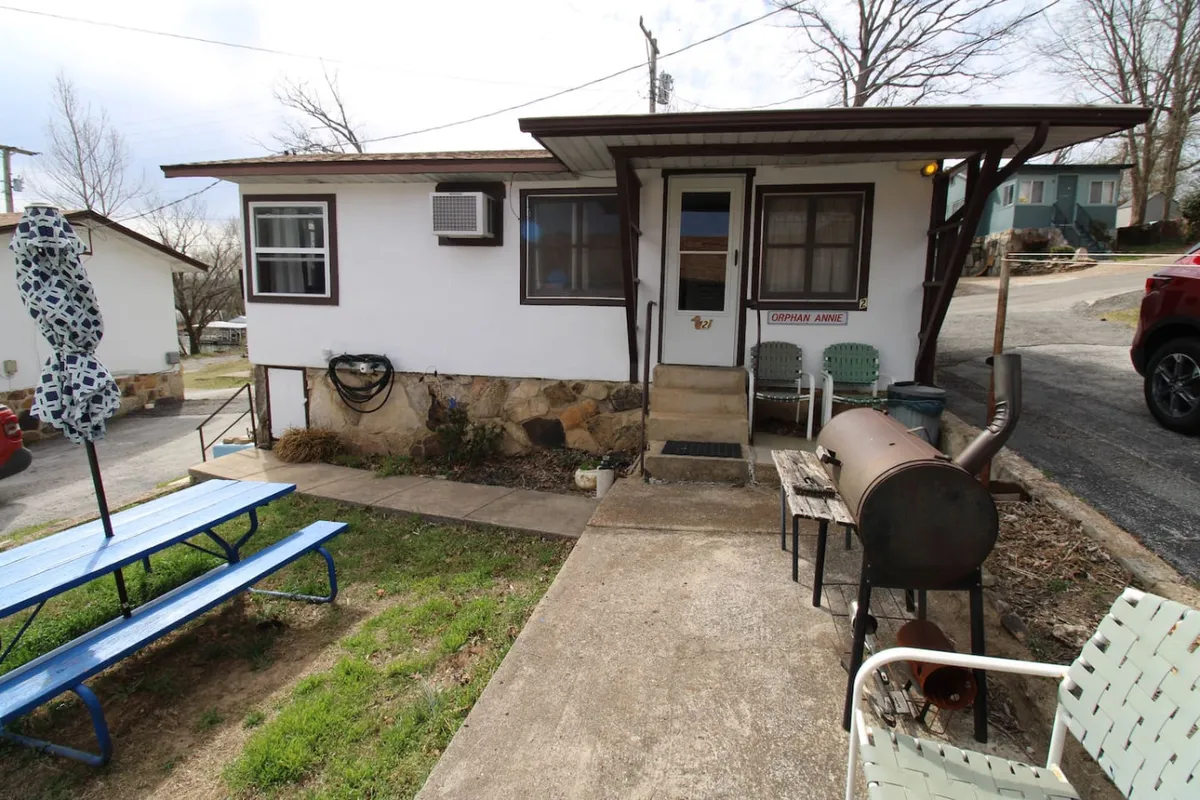 An outdoor area with a blue picnic table, a smoker grill, and a rock wall near the entrance door. The setup is casual and welcoming, ideal for barbecuing, dining outside, or relaxing with friends and family. The warm evening light adds to the inviting mood.