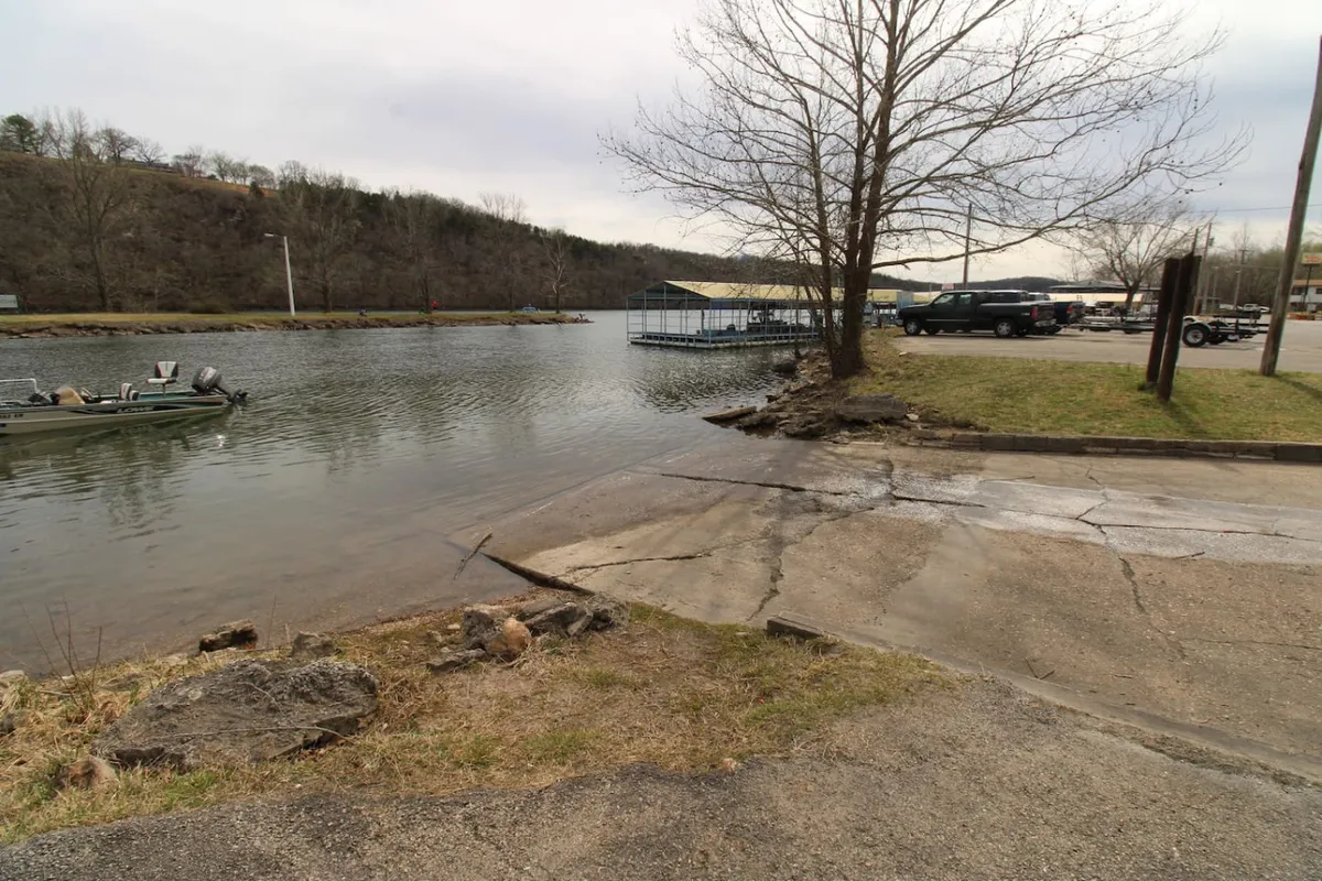 A tranquil lakeside view featuring a calm water surface, a boat docked at the shore, a bare tree, and a rocky shoreline. The scene is perfect for fishing, boating, or sharing a peaceful picnic with friends or family.