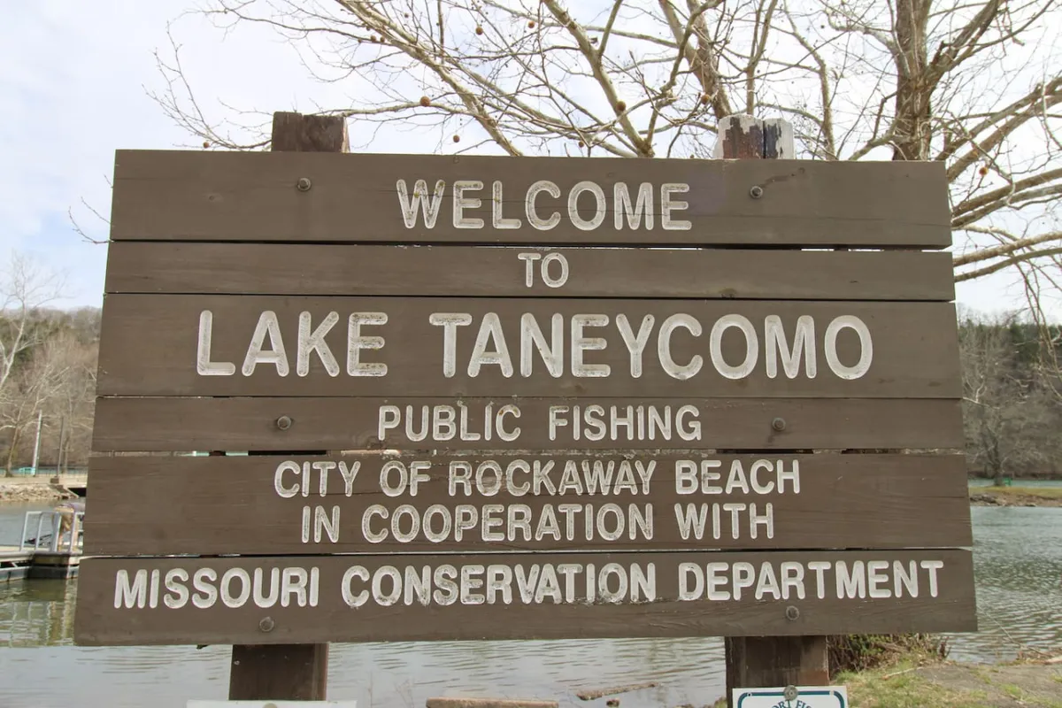 The image shows a cozy cabin exterior nestled among trees on the peaceful shores of Lake Taneycomo. A welcoming sign greets guests at the entrance, while the tranquil lake stretches out with gentle hills in the distance. The setting is calm, with sunlight filtering through the trees, perfect for both sunrise fishing and relaxing by the water.