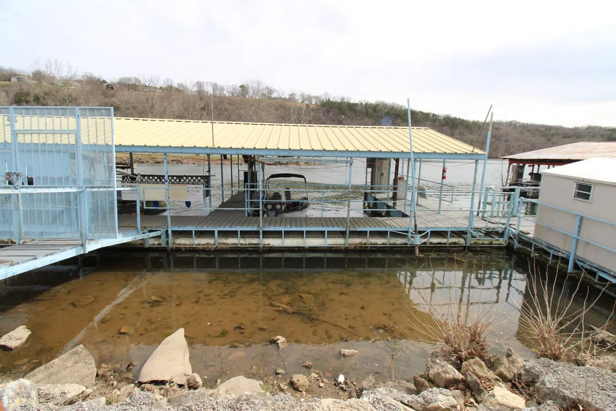 A peaceful lakeside dock with a yellow-roofed structure, boats moored alongside, and calm water reflecting the early morning or sunset light. People are seen preparing for boating or fishing, and the area is set up for relaxing by the water or gathering with friends.