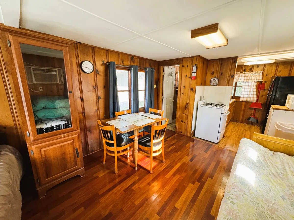 A homey interior scene displaying a dining table and chairs, a wooden cabinet, visible kitchen appliances, and a sofa. The arrangement creates an inviting atmosphere, ideal for family meals, game nights, and enjoying the company of loved ones in a lakeside retreat.