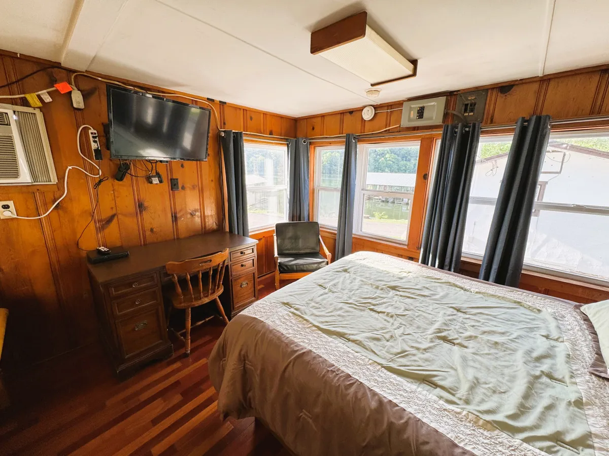 A comfortable bedroom featuring a cozy bed positioned near large windows that let in natural light. There's a small desk for writing or working, and a television mounted on the wall for evening relaxation. The space is uncluttered and peaceful, inviting restful sleep and quiet moments. The overall decor is simple and designed for comfort, ideal for couples or solo travelers seeking tranquility.