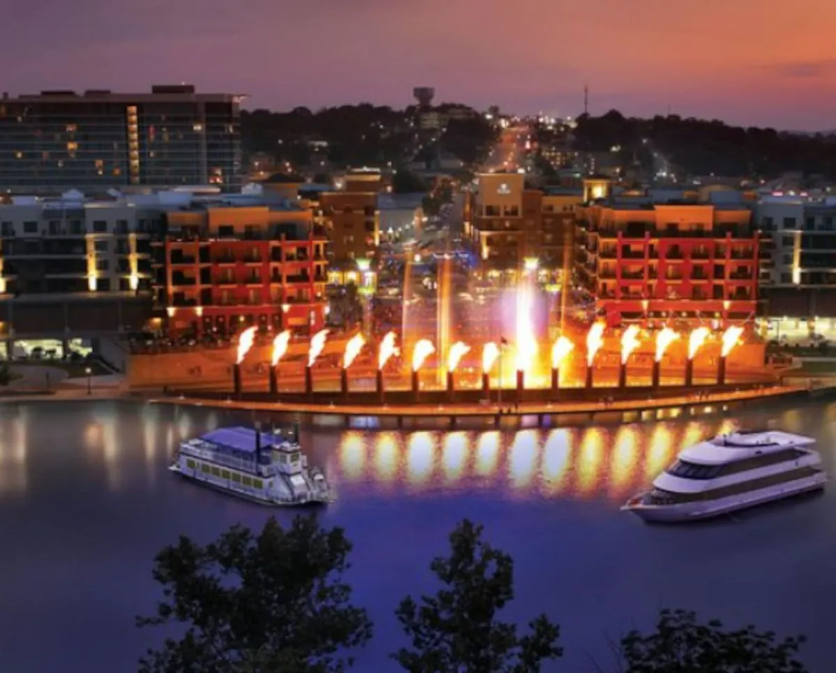 A vibrant nighttime waterfront scene with fountains alight with flames, boats gently floating on the water, and colorful buildings with twinkling lights along the promenade. People can be seen dining at waterfront restaurants and enjoying the lively atmosphere.