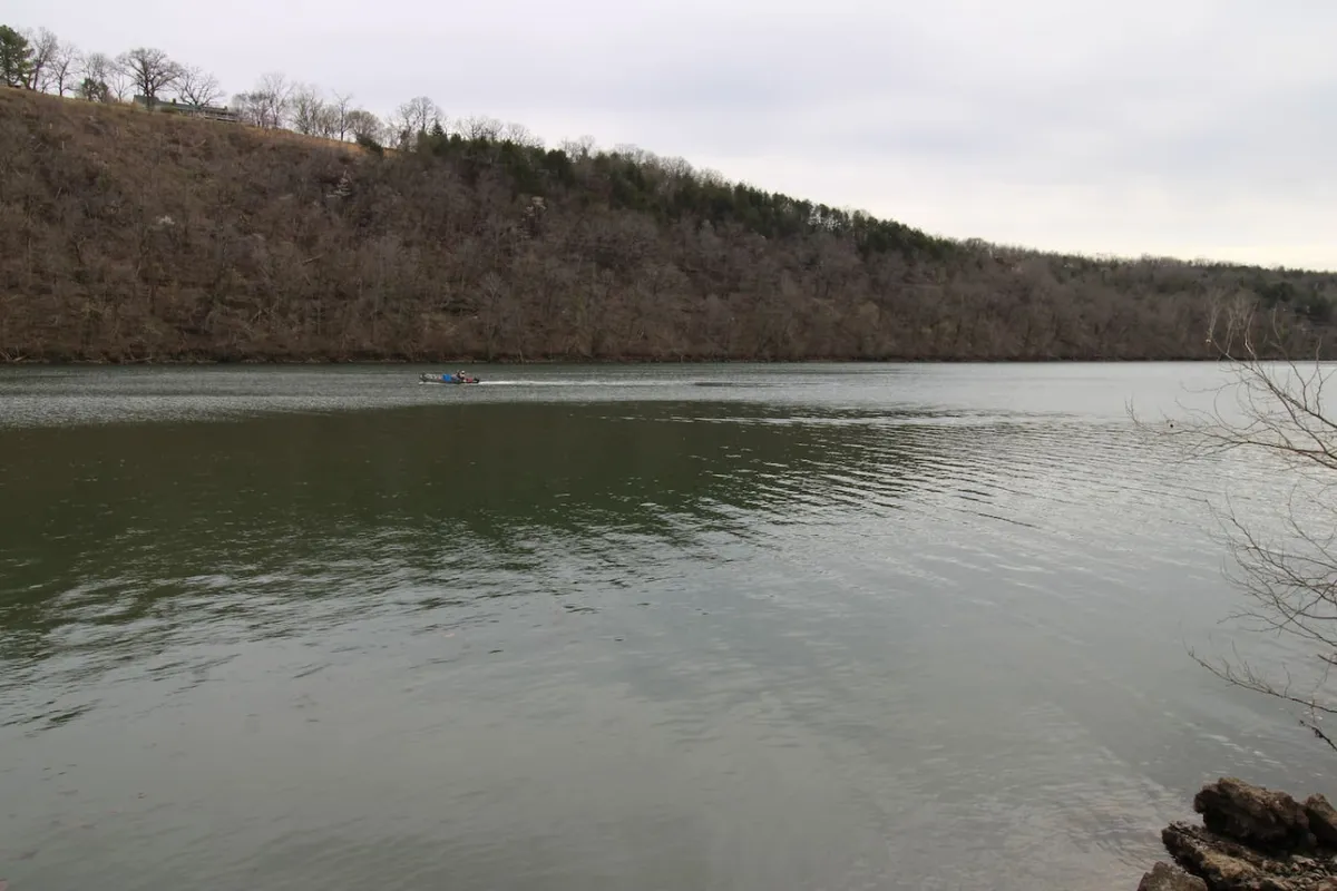 A tranquil view of Lake Taneycomo’s calm surface, with a distant boat on the water and a tree-lined shore creating a peaceful natural setting. The scene suggests early morning or late afternoon, ideal times for fishing or boating.
