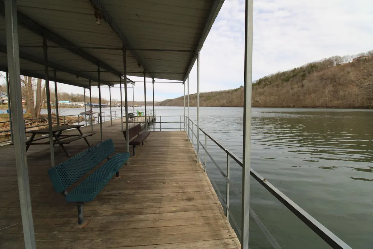 A peaceful lakeside scene featuring a wooden dock with benches, overlooking a calm water surface and gently rolling hills in the background. The setting invites guests to enjoy activities like fishing, picnicking, boating, or simply relaxing by the water.