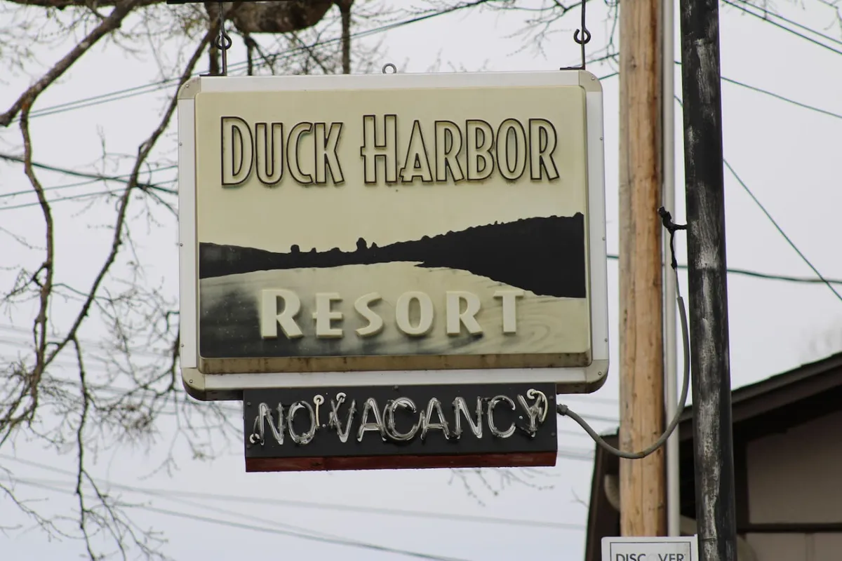 A welcoming exterior shot featuring the Duck Harbor sign and a glimpse of the lakeside resort, with the water visible beyond. The mood is tranquil, and the sign suggests a friendly, inviting atmosphere for visitors.